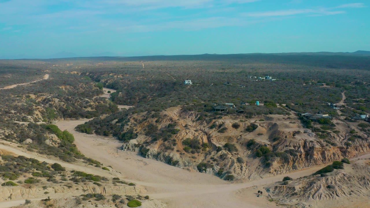 Aerial frame at Shipwrecks Beach, Baja California Sur, showing a pale dirt road junction through eroded brown terrain, sparse brush, distant hills, and no visible people.