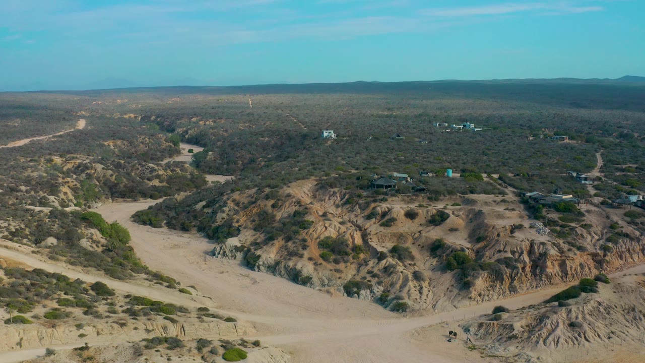 Aerial frame at Shipwrecks Beach, Baja California Sur, showing dry road tracks crossing sandy ground, eroded slopes in the foreground, dark scrubland beyond, and no visible people.