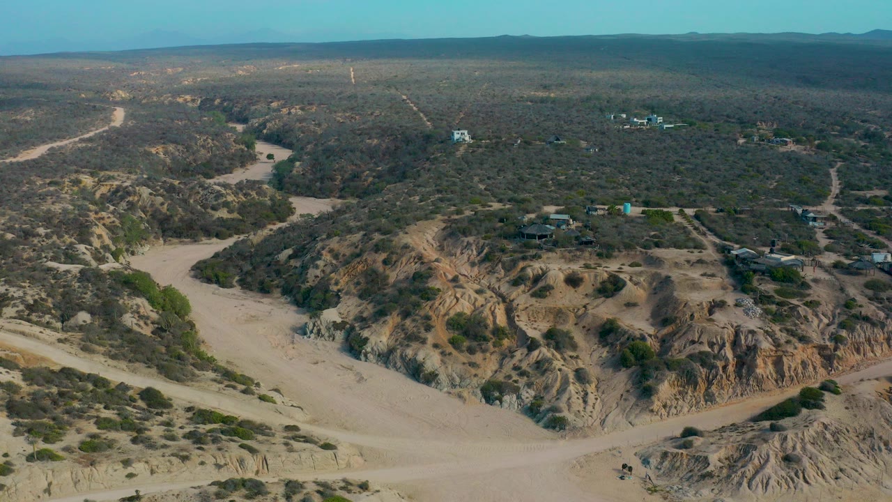 Aerial frame from Shipwrecks Beach, Baja California Sur, Mexico, showing a tan dry wash below a rocky ridge, scattered scrub, distant dirt tracks, and no visible people.
