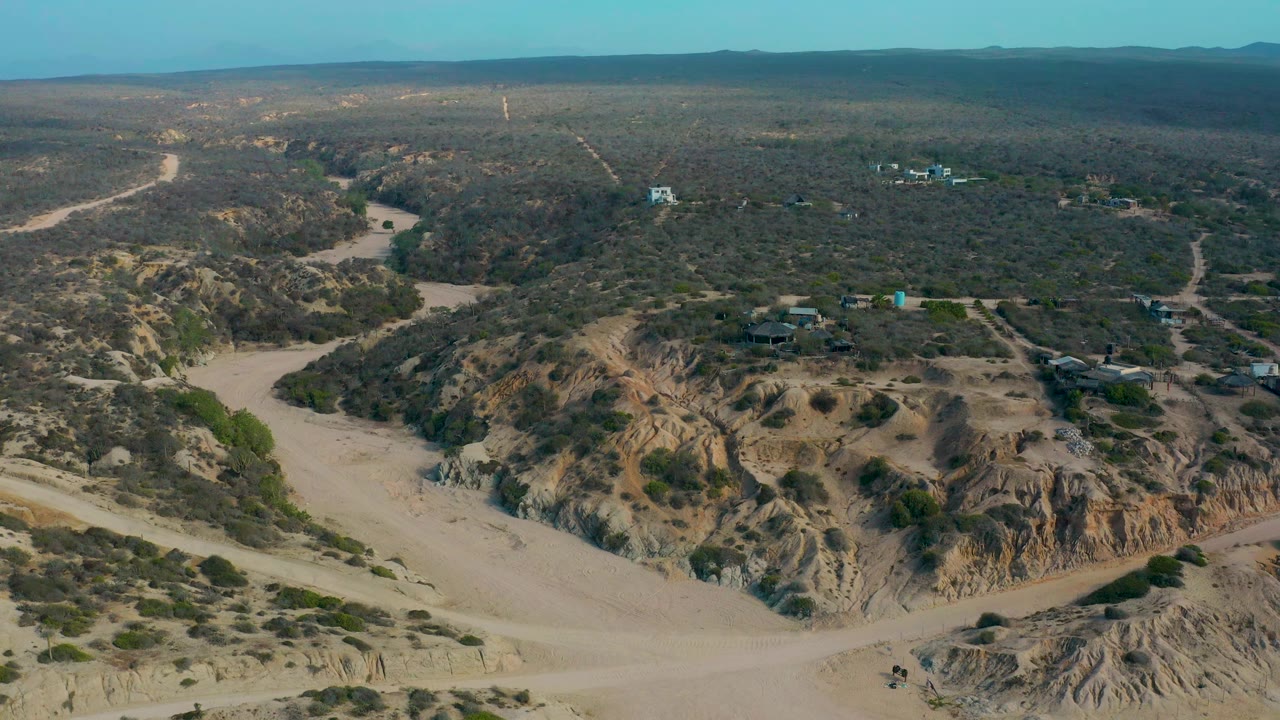 Wide aerial frame at Shipwrecks Beach in Baja California Sur, Mexico, with a pale dirt channel curving past rocky ground, scrub-covered flats, distant buildings, and no visible people.
