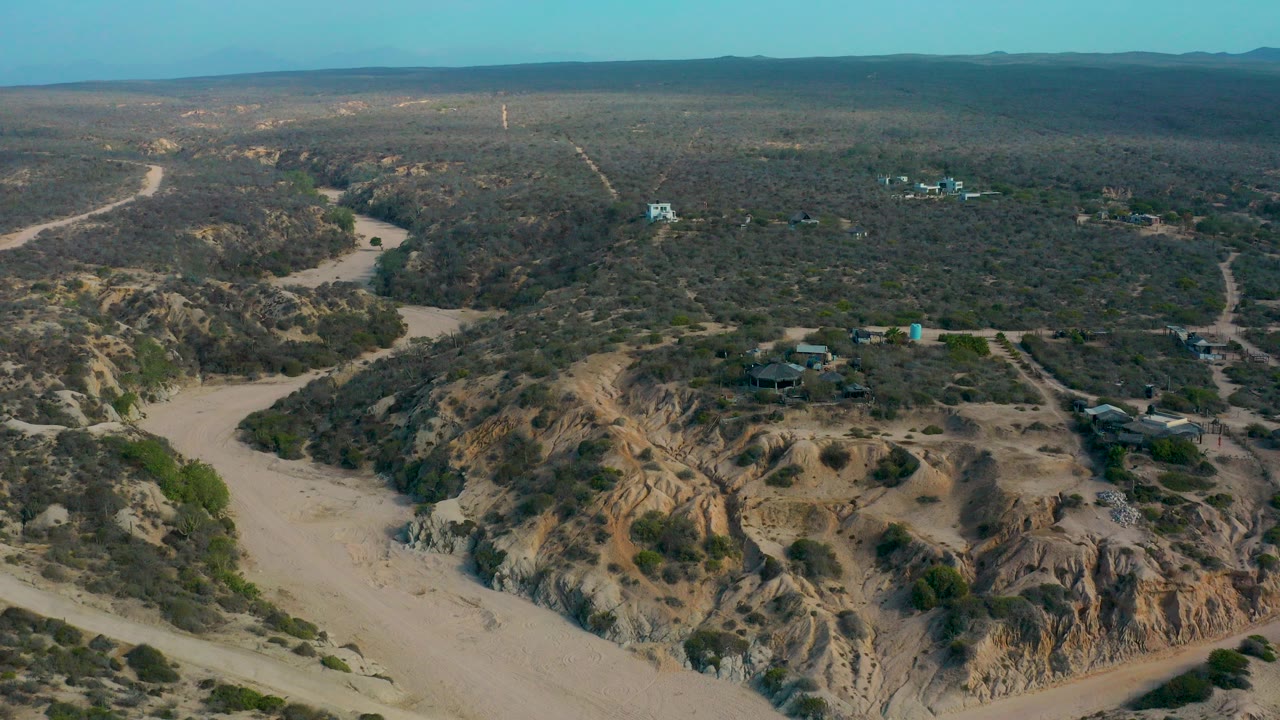 Aerial frame over Shipwrecks Beach, Baja California Sur showing a sandy wash, scrub-covered ridges, dirt trails, scattered small buildings, distant hills, and no visible people.