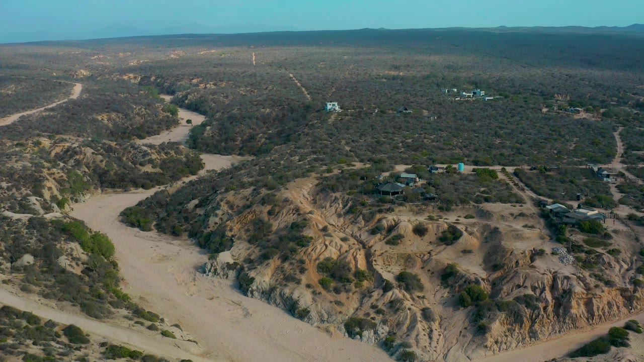 Aerial view at Shipwrecks Beach, Baja California Sur with a pale sandy wash curving through green scrub, low eroded slopes, dirt roads, small structures, and no visible people.