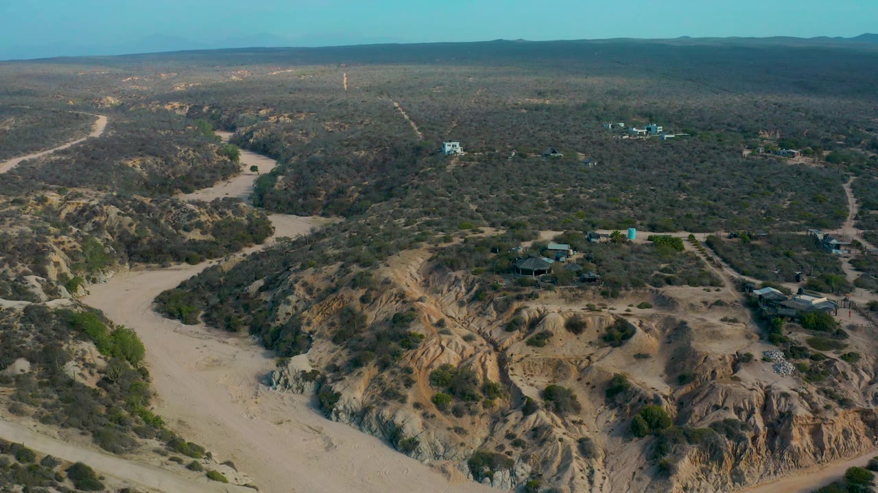 Wide aerial frame over Shipwrecks Beach, Baja California Sur showing sandy canyon ground in the foreground, scattered buildings among scrub in the midground, distant low hills, and no visible people.
