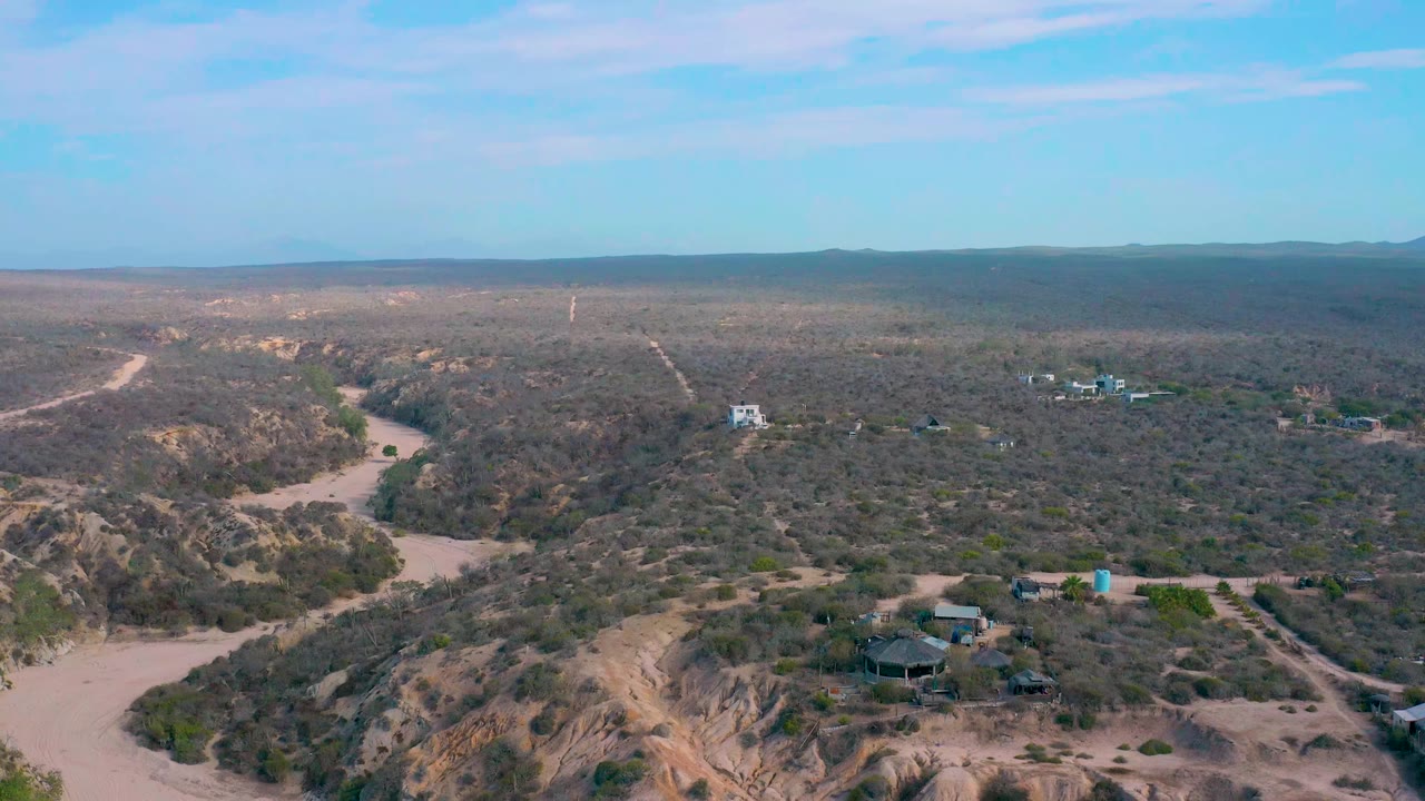 A second aerial view at Shipwrecks Beach shows the eroded dune ridge lower in frame, a pale wash curving along the left side, scattered buildings beyond the scrub, and no visible people.