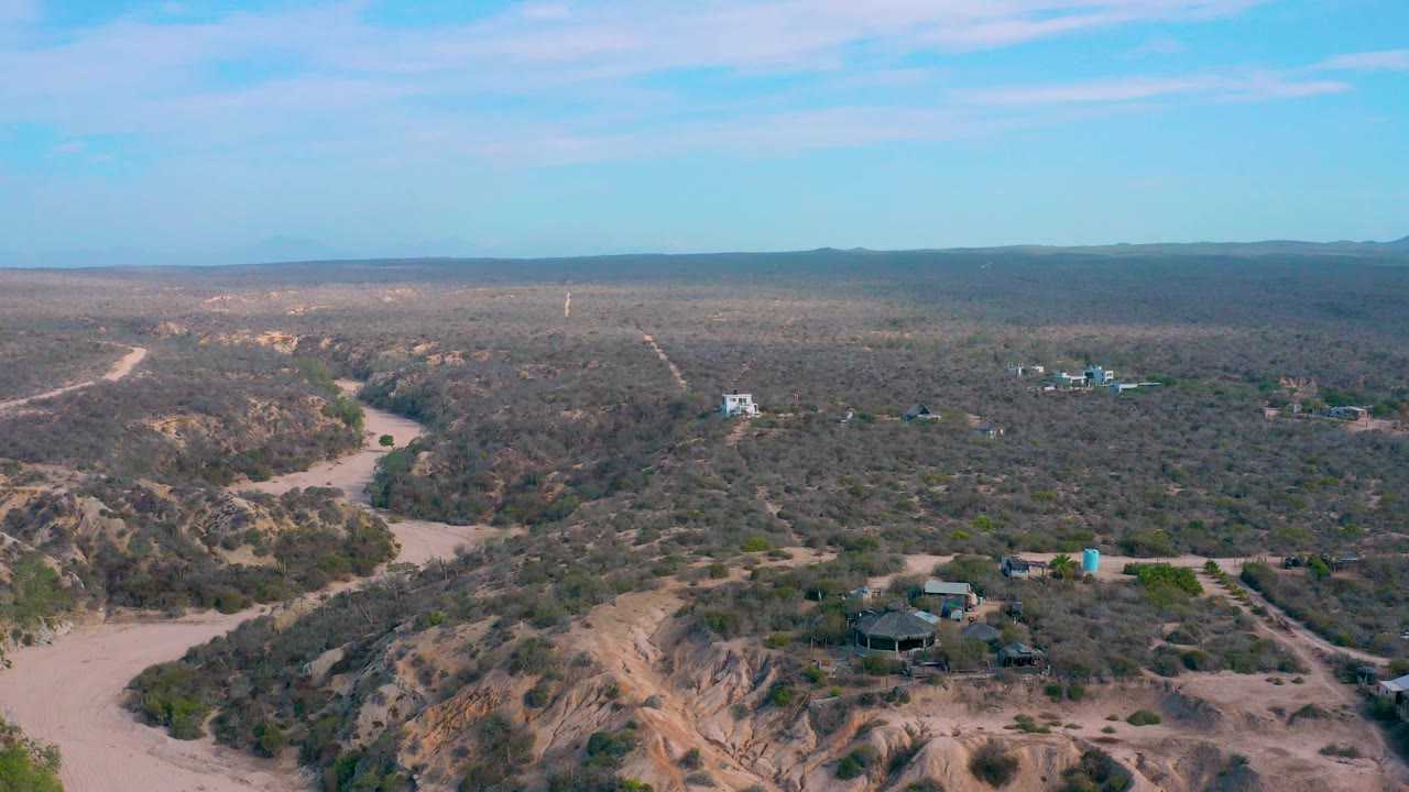 The frame looks across Shipwrecks Beach dune terrain with tan ridges in the foreground, scrub and dirt routes across the middle distance, a small cluster of buildings to the right, and no visible people.