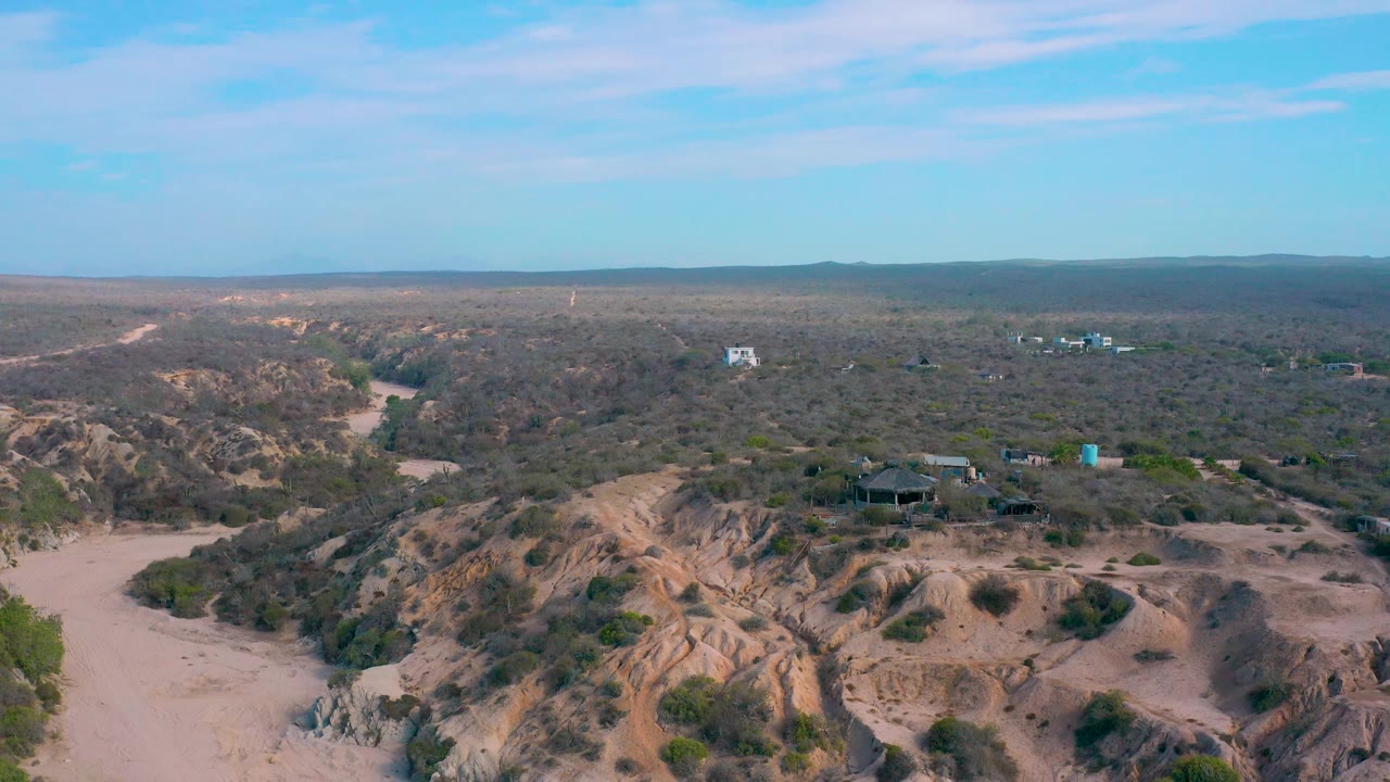 Aerial frame from Shipwrecks Beach, Baja California Sur showing eroded tan hills across the foreground, scrubland beyond, small distant structures, and no visible people.