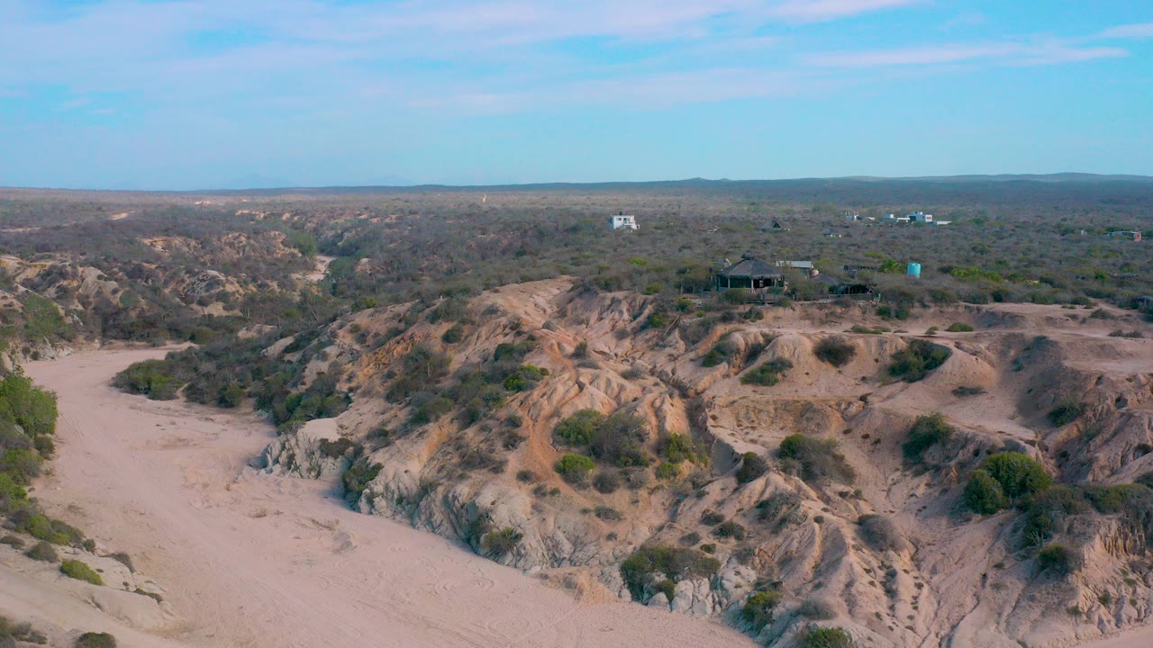 Aerial frame at Shipwrecks Beach, Baja California Sur, showing pale sand, eroded ridges, scattered shrubs, distant structures, and no visible people.