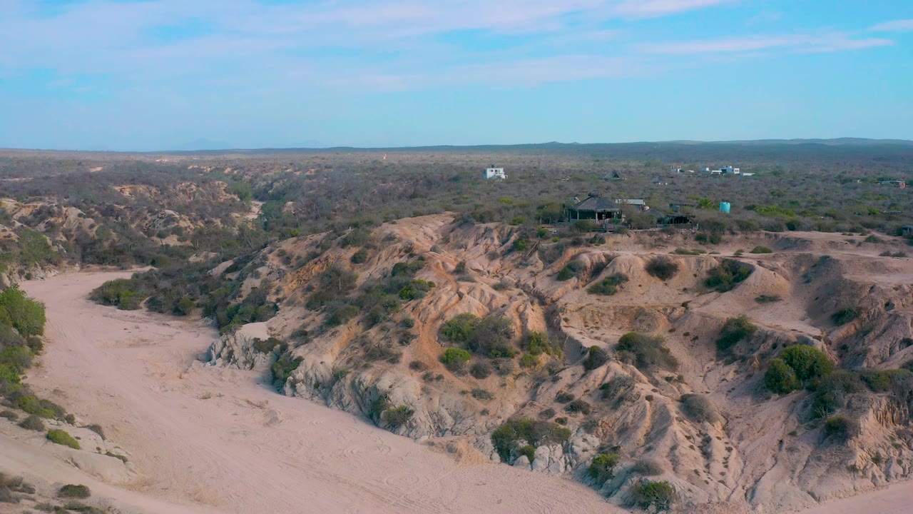 High aerial frame from Shipwrecks Beach showing the sandy road curving through dry terrain, with eroded tan ridges, sparse shrubs, small distant structures, and no visible people.