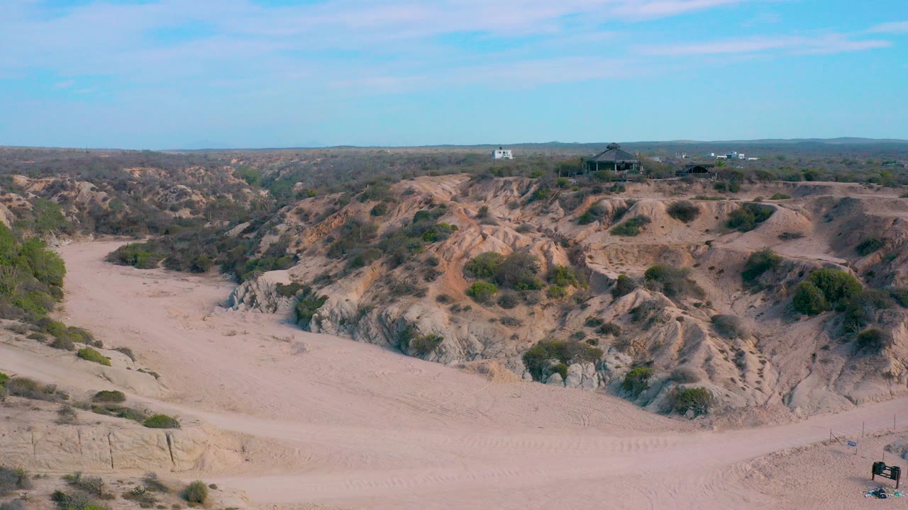 Wide aerial view at Shipwrecks Beach in Baja California Sur showing pale sand, eroded scrub-covered hills, a distant ridge structure, and no visible people.