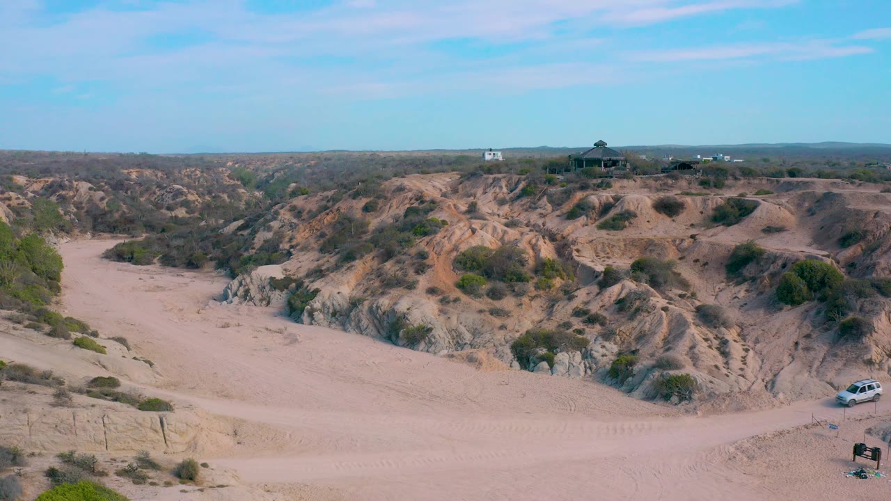 A white vehicle appears near the right side of the sandy terrain at Shipwrecks Beach in Baja California Sur, with scrub hills behind it and no visible people.