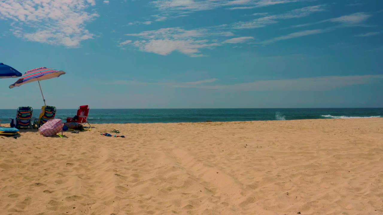 A blue umbrella and striped umbrella shade beach chairs on Shipwrecks Beach, with scattered items on the sand and a calm ocean horizon in the background.