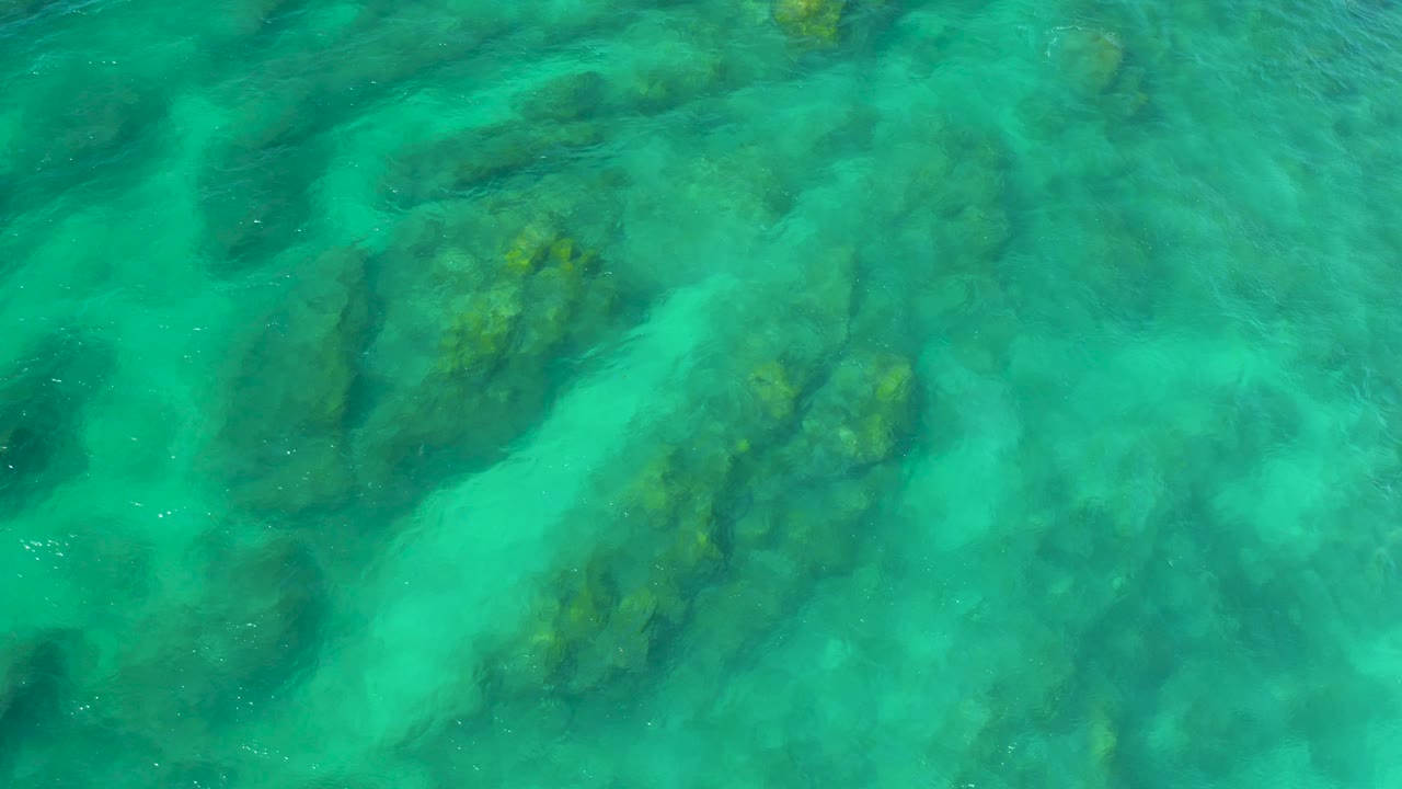 A straight-down view at Shipwrecks Beach, Baja California Sur, shows shallow turquoise water, darker reef ridges, cloudy pale sand, and no visible people.