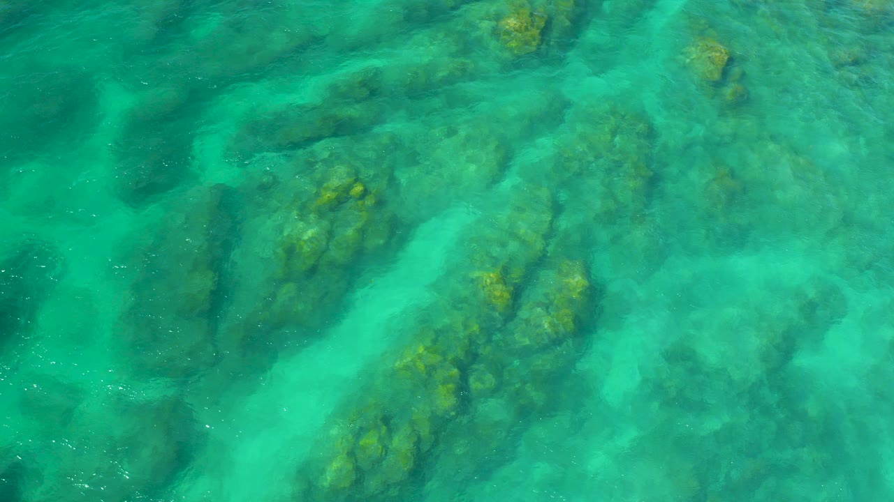 Top-down aerial view of turquoise water and dark submerged rock formations at Shipwrecks Beach, Baja California Sur, with no visible people.