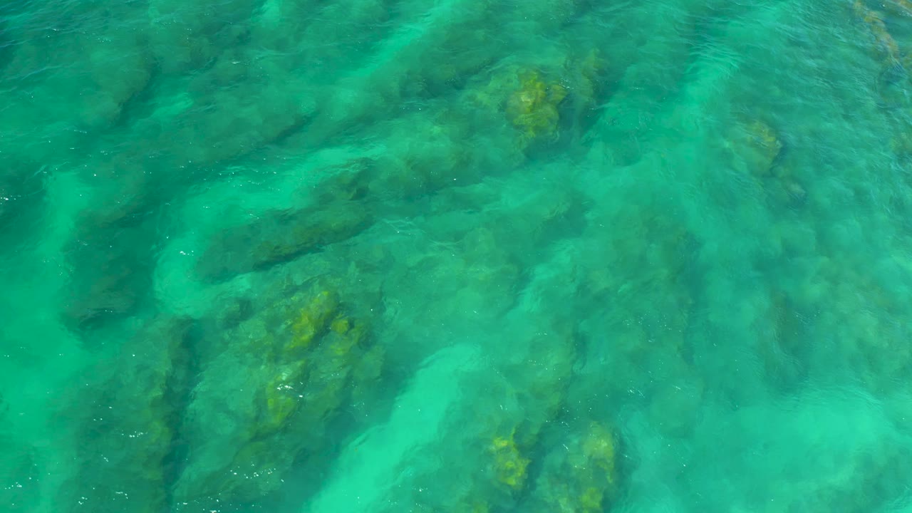 Bright turquoise water reveals clustered submerged rocks and faint surface texture at Shipwrecks Beach, Baja California Sur, with no visible people.