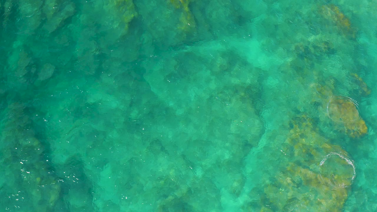 Overhead view of clear aqua water at Shipwrecks Beach, Baja California Sur, with darker submerged reef shapes and no visible people.