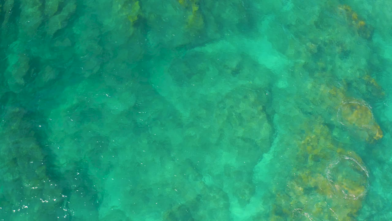 Clear aqua ocean water reveals amber-green reef patches and pale sandy areas below at Shipwrecks Beach, Baja California Sur, with no visible people.