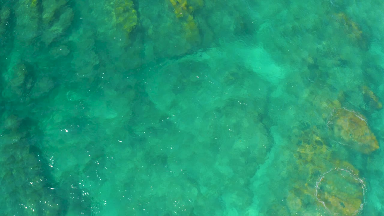 Aerial-looking overhead composition of clear water and dark reef bands at Shipwrecks Beach, with small surface ripples and no visible people.
