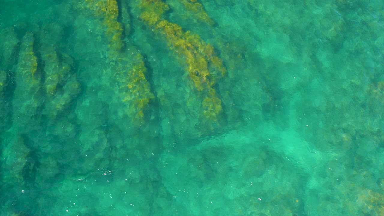 Overhead view of turquoise seawater at Shipwrecks Beach, Baja California Sur, with dark submerged rock shapes and yellow-green plant growth, no visible people.