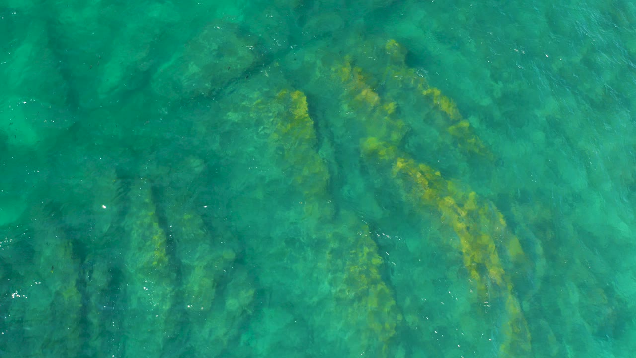 Straight-down view of turquoise water over dark submerged rock ridges at Shipwrecks Beach, Baja California Sur, with faint surface ripples and no visible people.