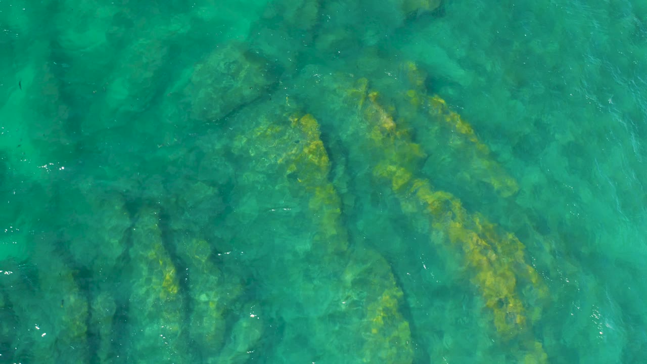 Teal water reveals darker submerged rock bands and yellow-green growth at Shipwrecks Beach, Baja California Sur, with no shore or people visible.