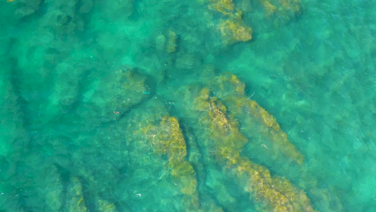 Top-down aerial frame of aqua ocean over elongated submerged rocks at Shipwrecks Beach, with faint surface ripples and no visible people.