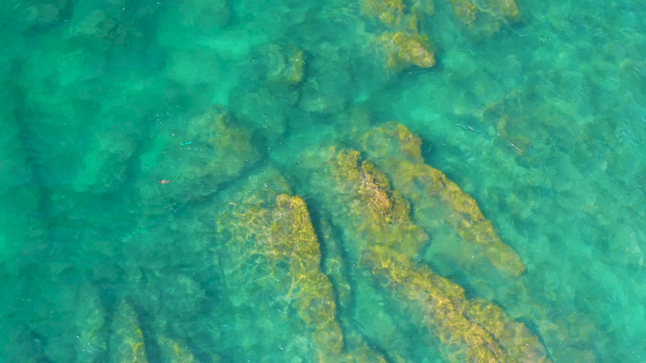 Straight-down aerial view of turquoise ocean with shallow rock forms below the surface at Shipwrecks Beach, and no visible people in frame.