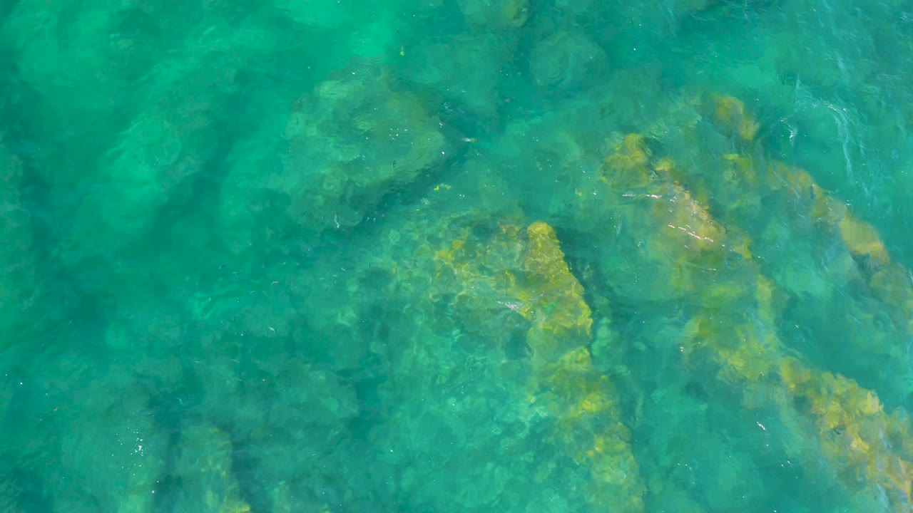 Aerial top-down frame of turquoise water at Shipwrecks Beach, where golden-brown submerged rock patches sit under the clear surface with no visible people.