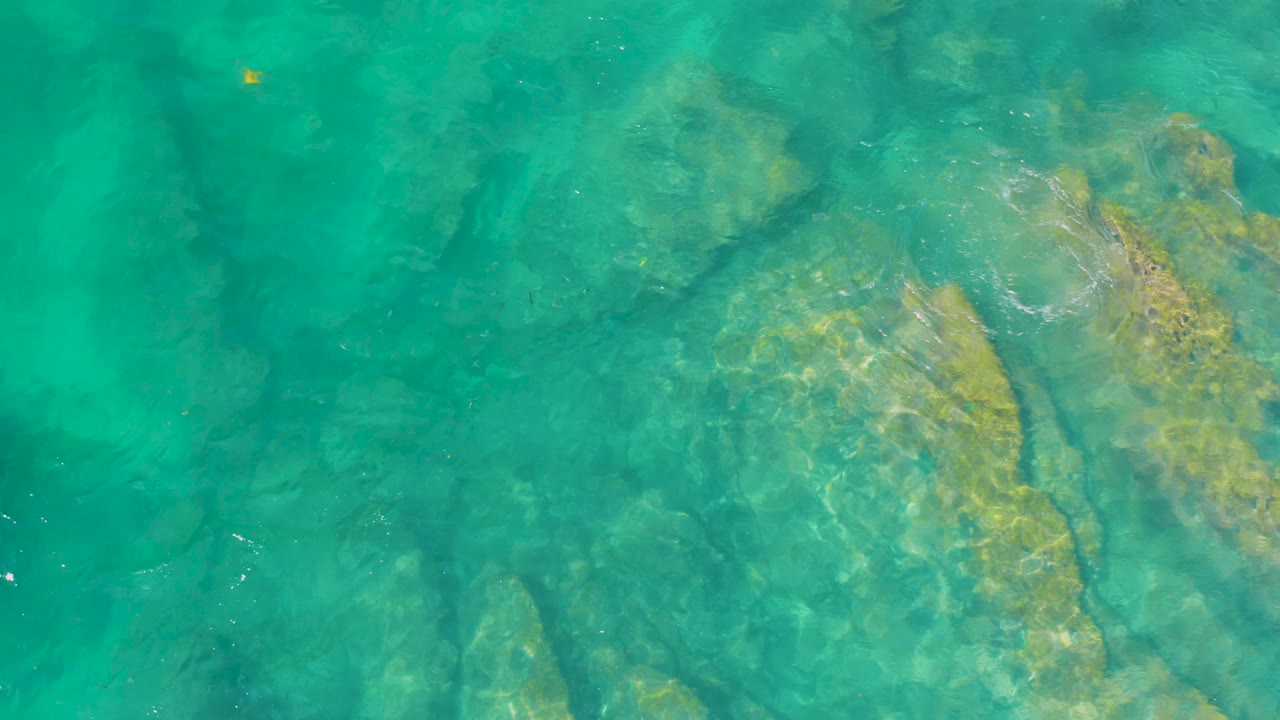 Green-blue water reveals a rocky seafloor at Shipwrecks Beach, with yellow patches on the rocks and light surface shimmer, no visible people.