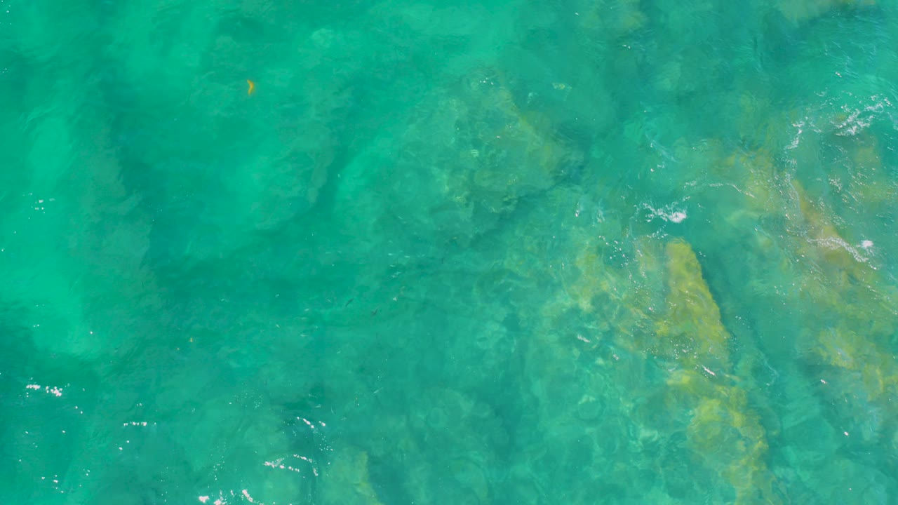 Top-down view of clear turquoise water over submerged beige rocks at Shipwrecks Beach, Baja California Sur, with faint ripples and no visible people.
