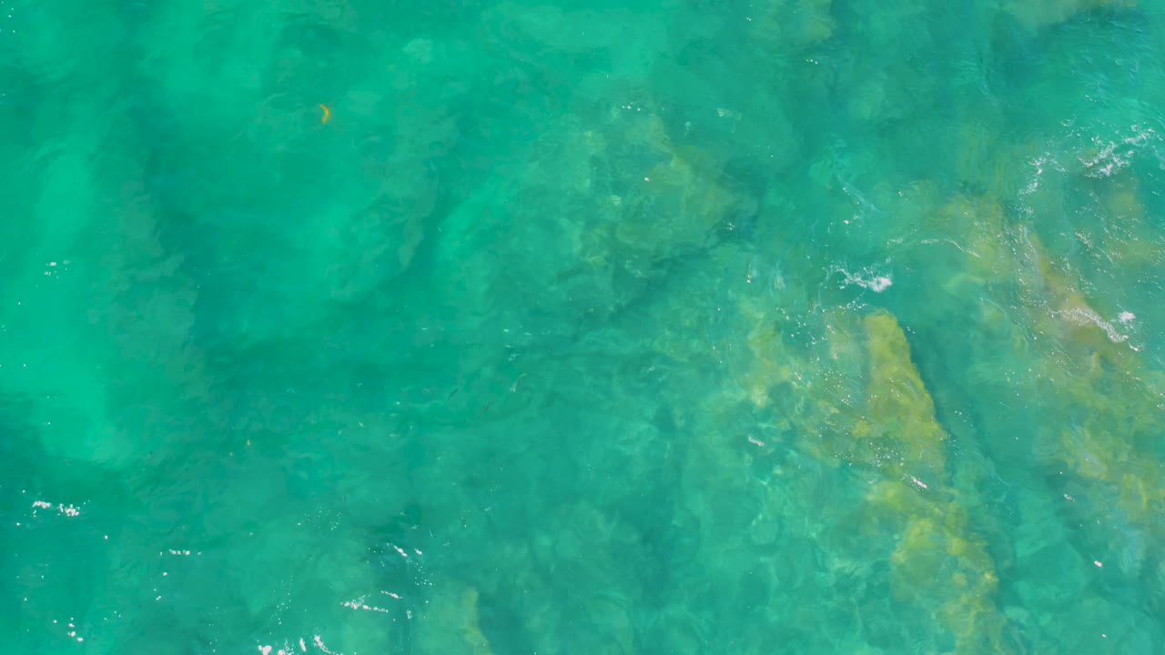 Blue-green water covers darker and beige rock shapes at Shipwrecks Beach, Baja California Sur, with small white glints on the rippled surface and no visible people.