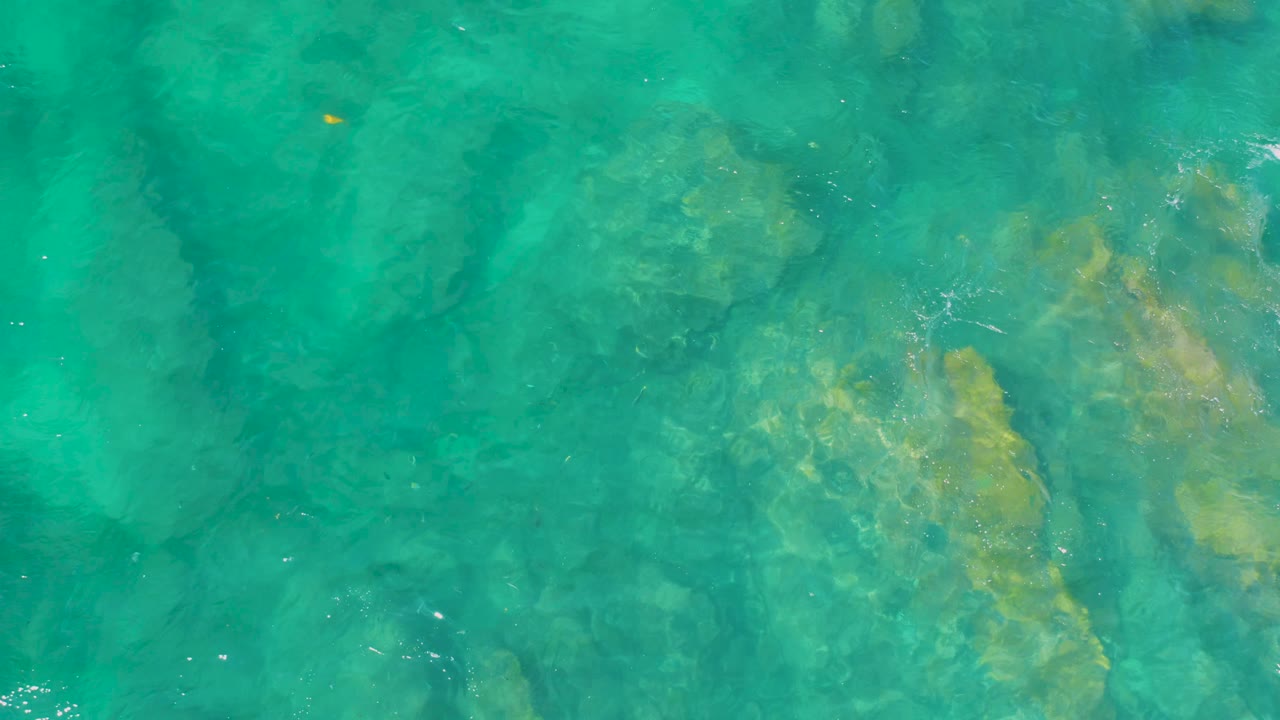 Turquoise water overlays uneven submerged rocks at Shipwrecks Beach, Baja California Sur, with a small orange speck near the upper area and no visible people.