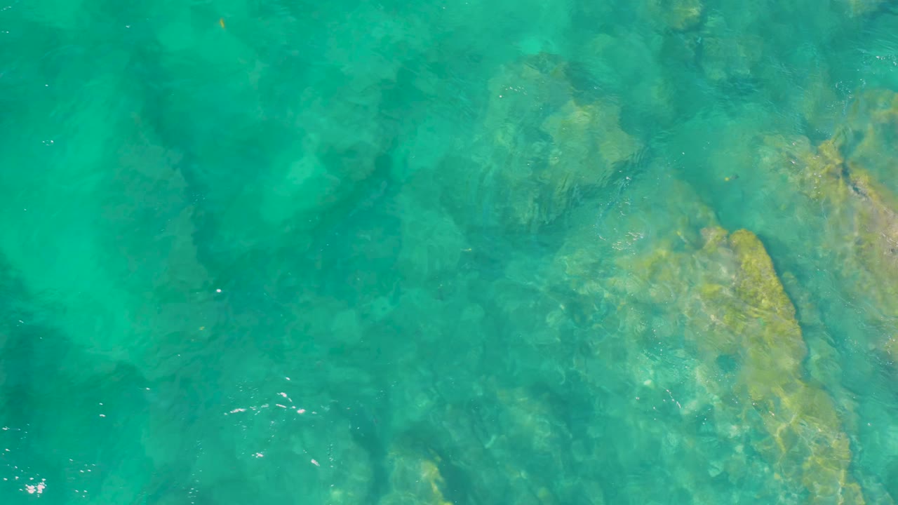 Blue-green ocean water passes over broad submerged rocks at Shipwrecks Beach, with small bright surface glints and no visible people.
