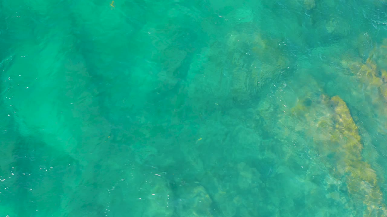 Rippling clear sea water reveals pale yellow-green rock patches beneath the surface at Shipwrecks Beach, with no visible people.