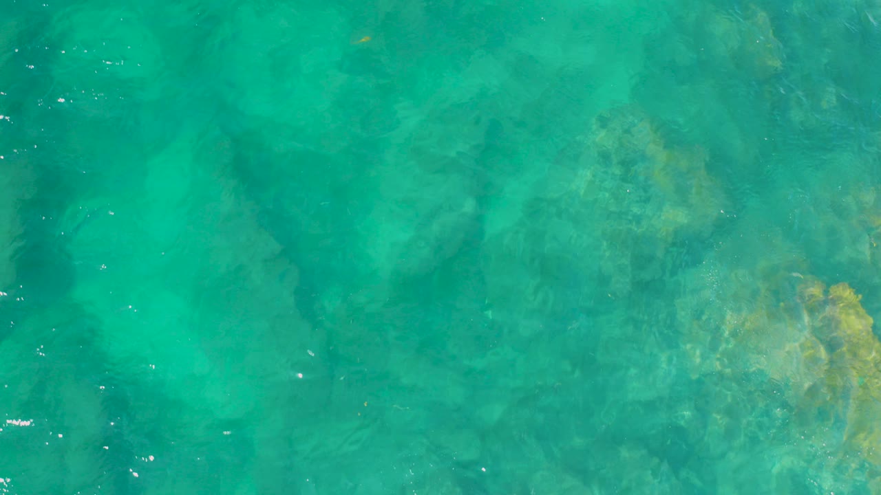 Overhead view of turquoise water at Shipwrecks Beach, Baja California Sur, with submerged rock shapes, pale sand, surface ripples, and no visible people.