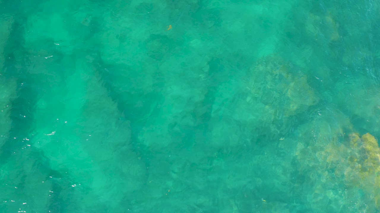 Top-down seawater view at Shipwrecks Beach shows a darker vertical rock shadow area, lighter sand below, rippled surface highlights, and no visible people.