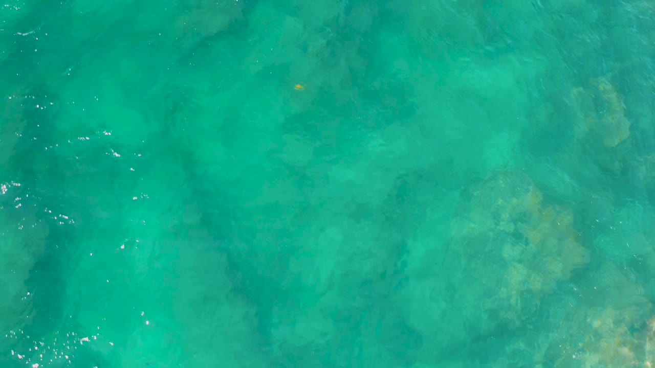 Overhead transparent water at Shipwrecks Beach, Baja California Sur, reveals yellow-green patches and darker underwater contours, with no visible people.