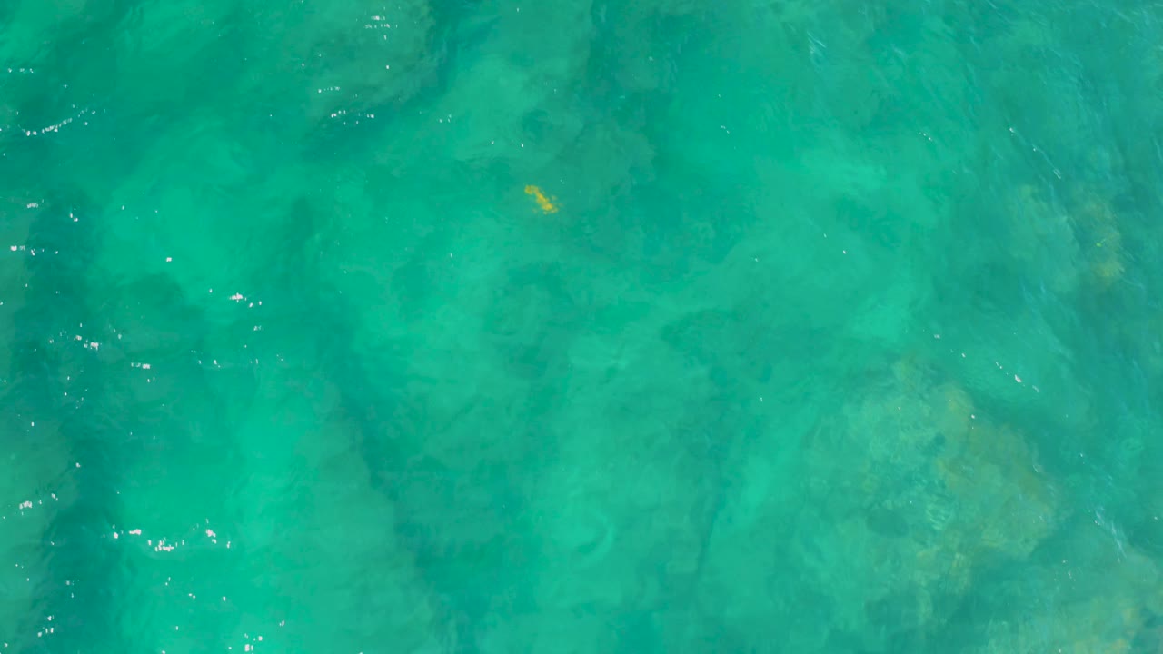 Turquoise water seen from above at Shipwrecks Beach, Baja California Sur, shows dark submerged bands crossing the frame and small bright sparkles on the surface, no visible people.