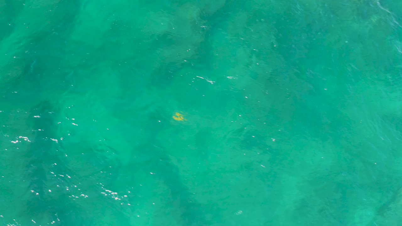 Overhead turquoise water at Shipwrecks Beach showing a small yellow speck against the sea surface, darker patches below, and no visible people.