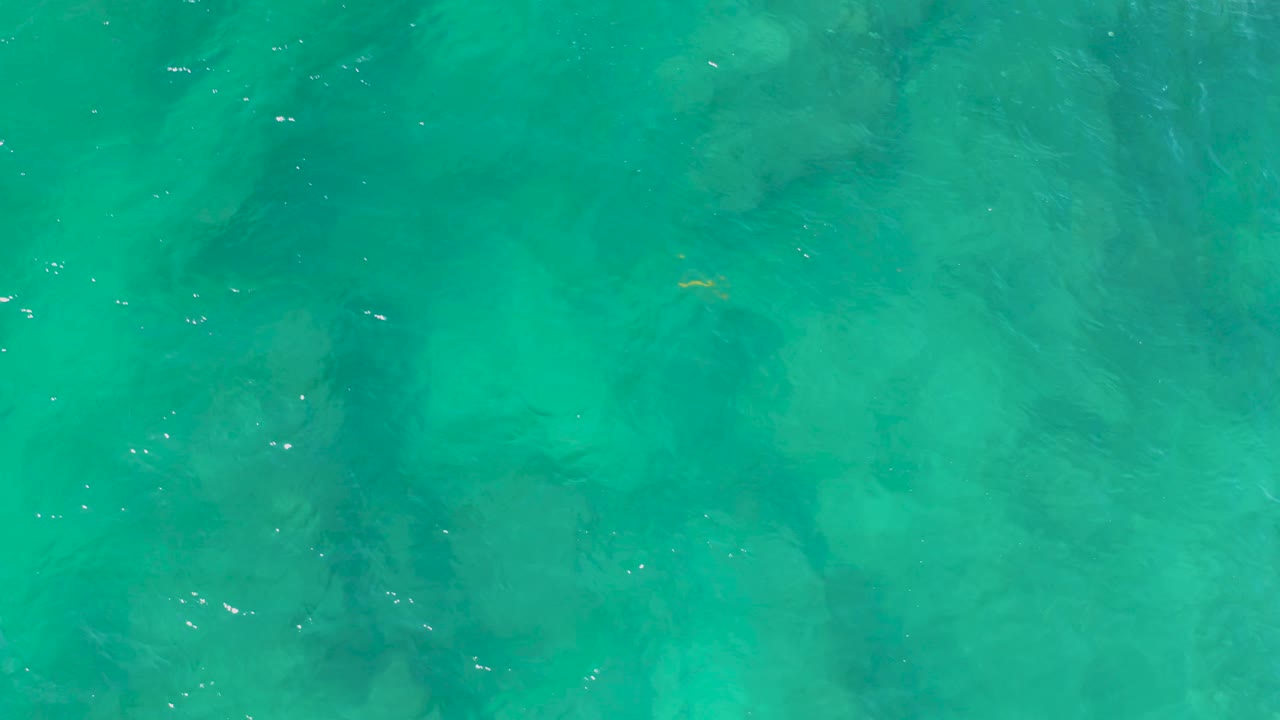 Overhead turquoise seawater at Shipwrecks Beach shows a small yellow fish-like shape near the lower center, dark submerged texture, and no visible people.