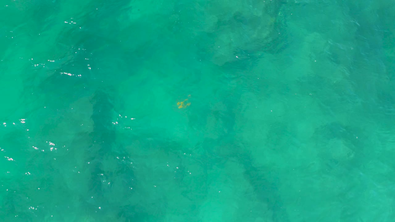 Overhead view of turquoise-green water at Shipwrecks Beach with faint dark shapes under or reflected on the surface and no visible people.