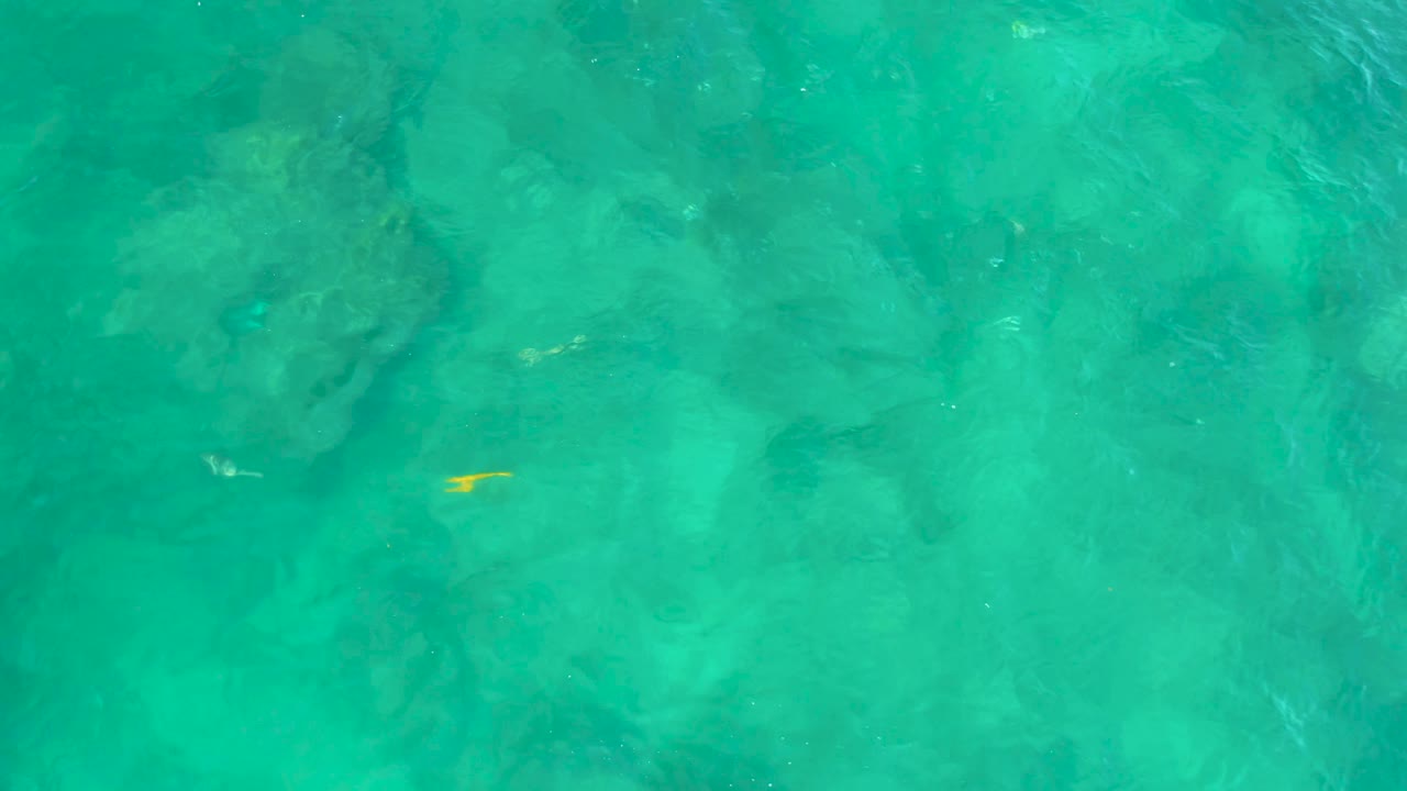 Rippling turquoise water fills the Shipwrecks Beach frame, with darker submerged forms below and only a faint yellow fish detail visible, no visible people.