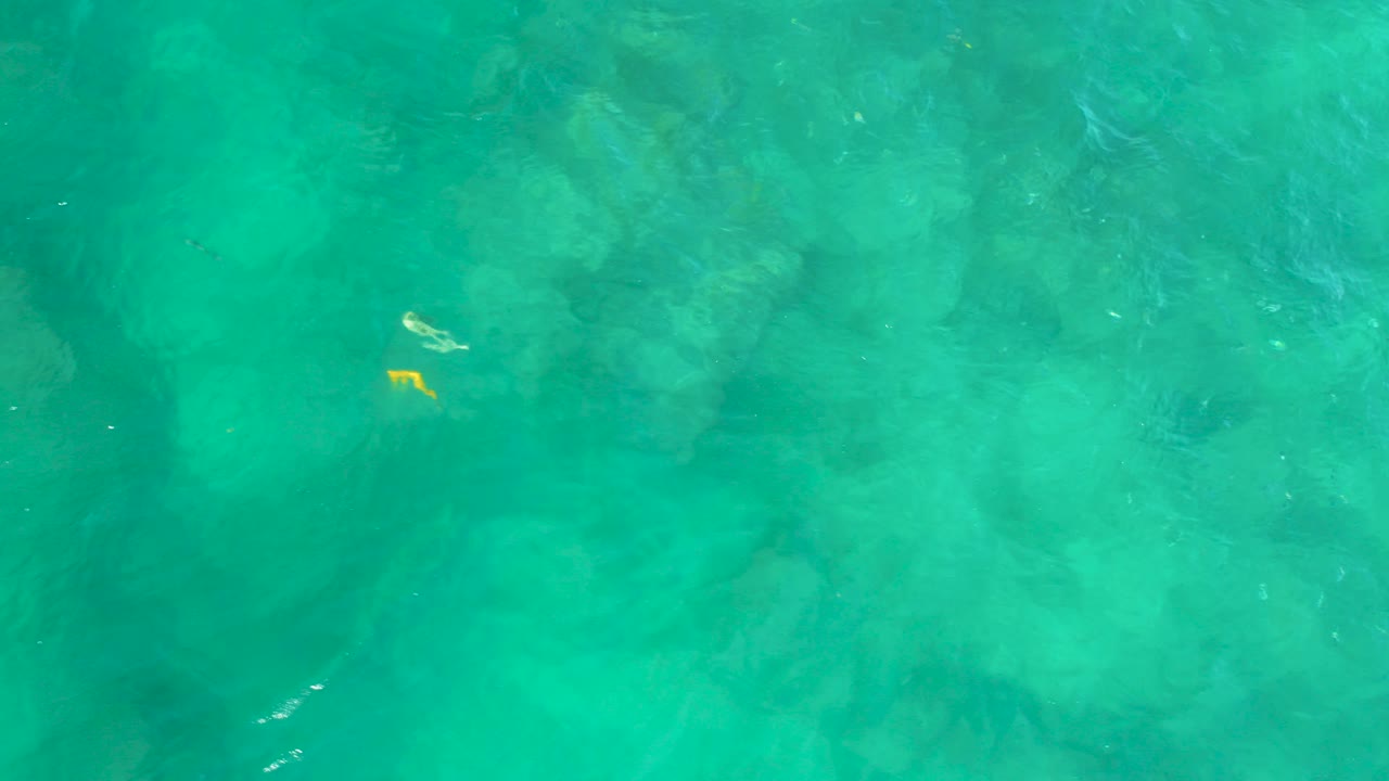 Overhead view of turquoise water at Shipwrecks Beach, Baja California Sur, with pale bottom texture, faint dark submerged shapes, small yellow flecks near the upper left, and no visible people.