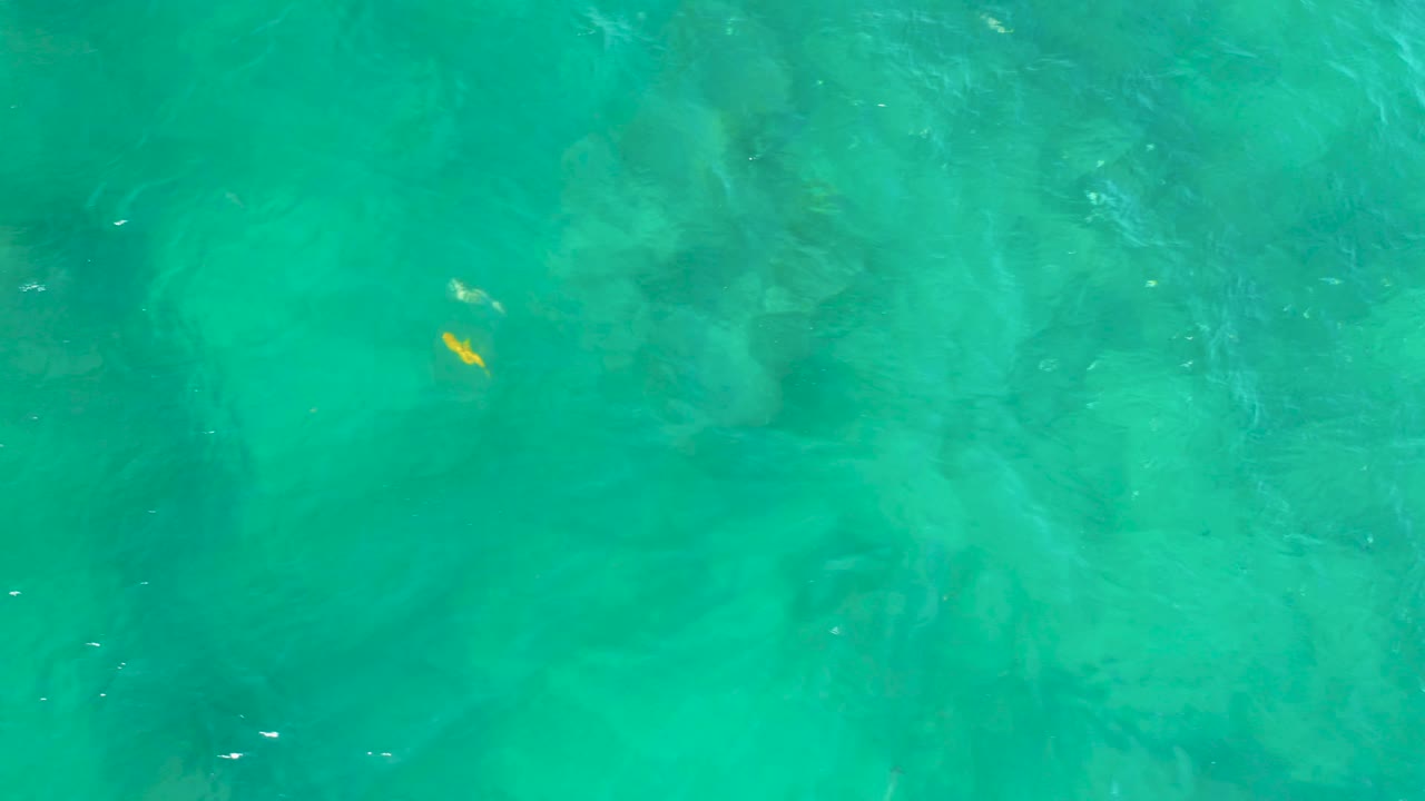 Top-down turquoise water at Shipwrecks Beach with cloudy pale bottom patterns, faint darker shapes beneath the surface, a small yellow fleck near the upper right, and no visible people.
