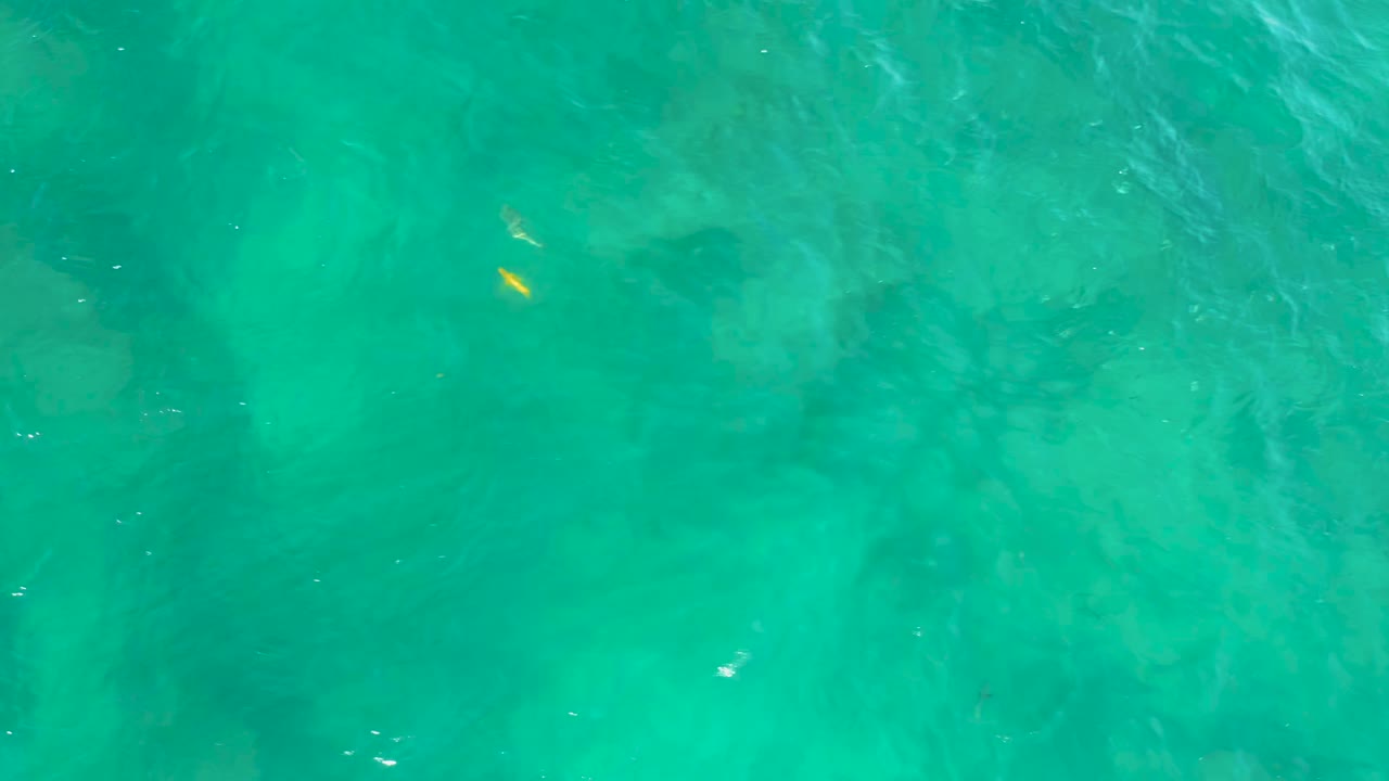 Overhead frame from Shipwrecks Beach showing rippled turquoise water, pale underwater texture, darker submerged areas, yellow flecks toward the left side, and no visible people.