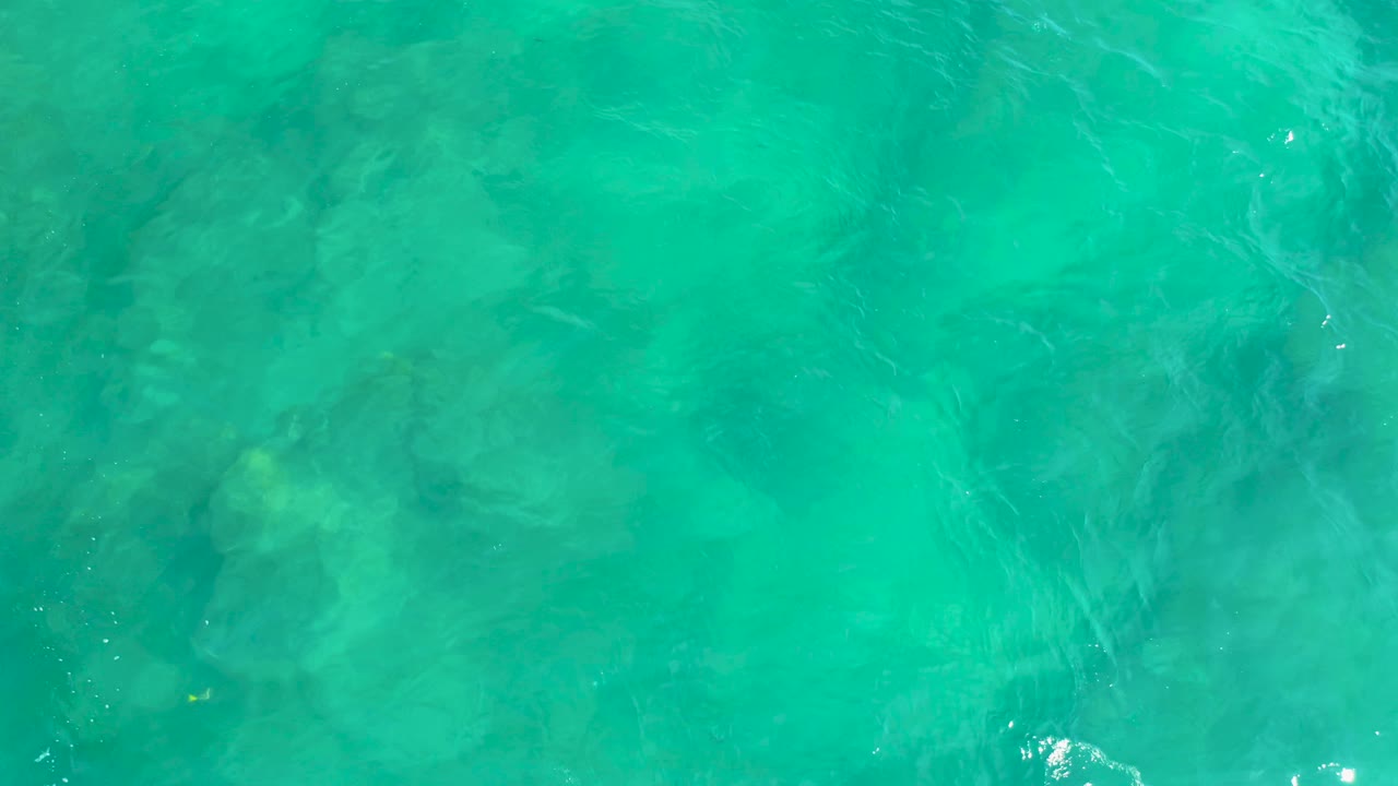 Top-down turquoise water at Shipwrecks Beach shows wavering surface light over darker underwater patches, with no visible people or objects above the water.