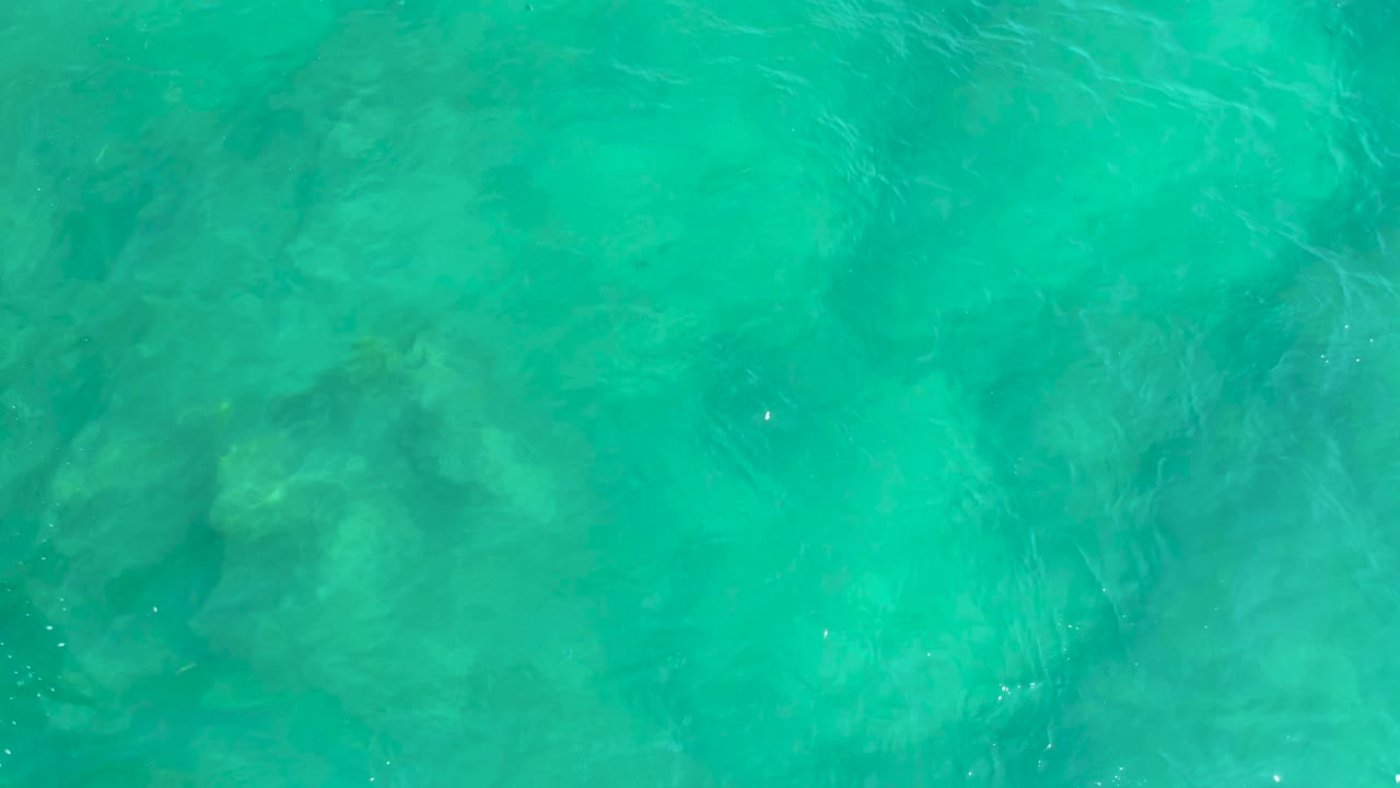 Overhead water at Shipwrecks Beach appears brighter near the center, with faint underwater rock-like shadows under the turquoise surface and no visible people.
