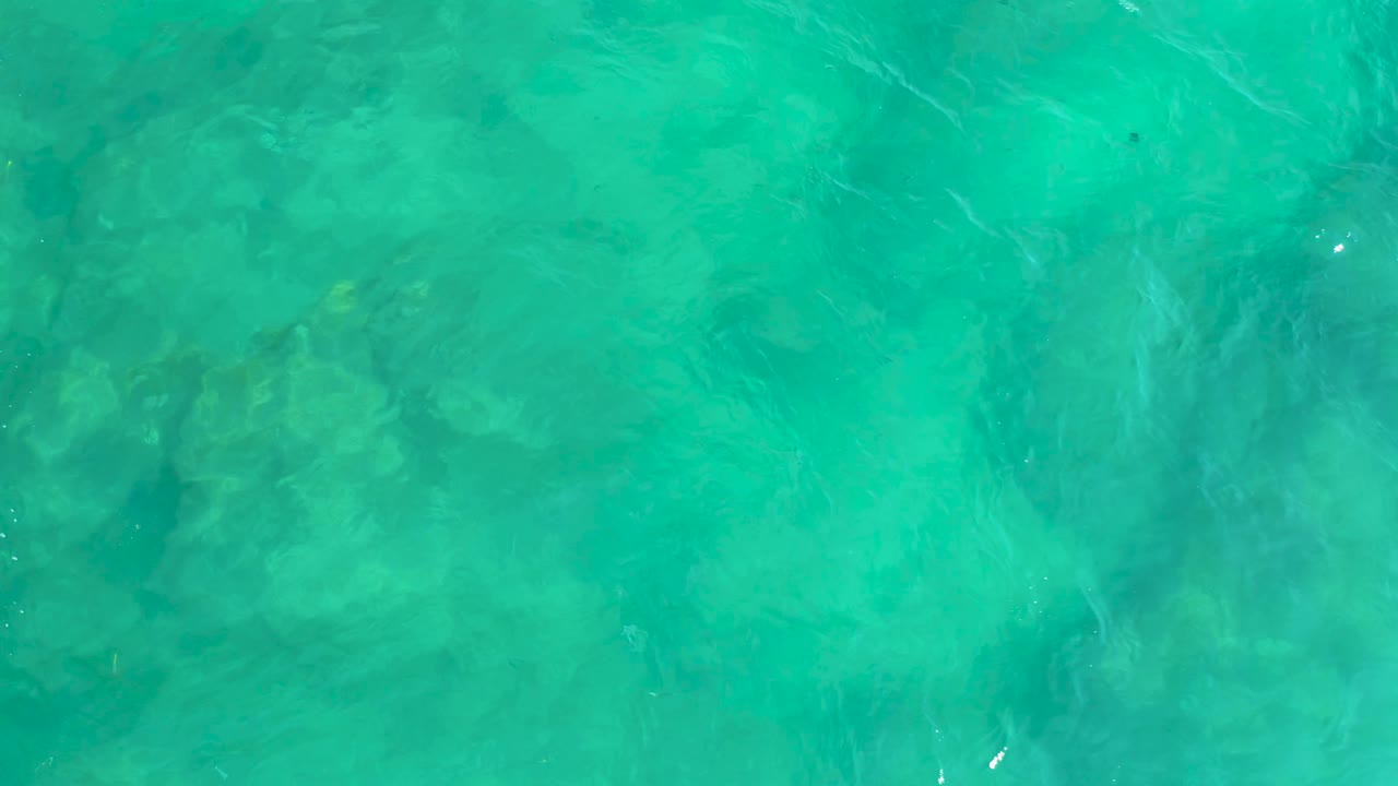 Blue-green water at Shipwrecks Beach in Baja California Sur, Mexico, with faint wave streaks crossing over darker submerged patches and no visible people.