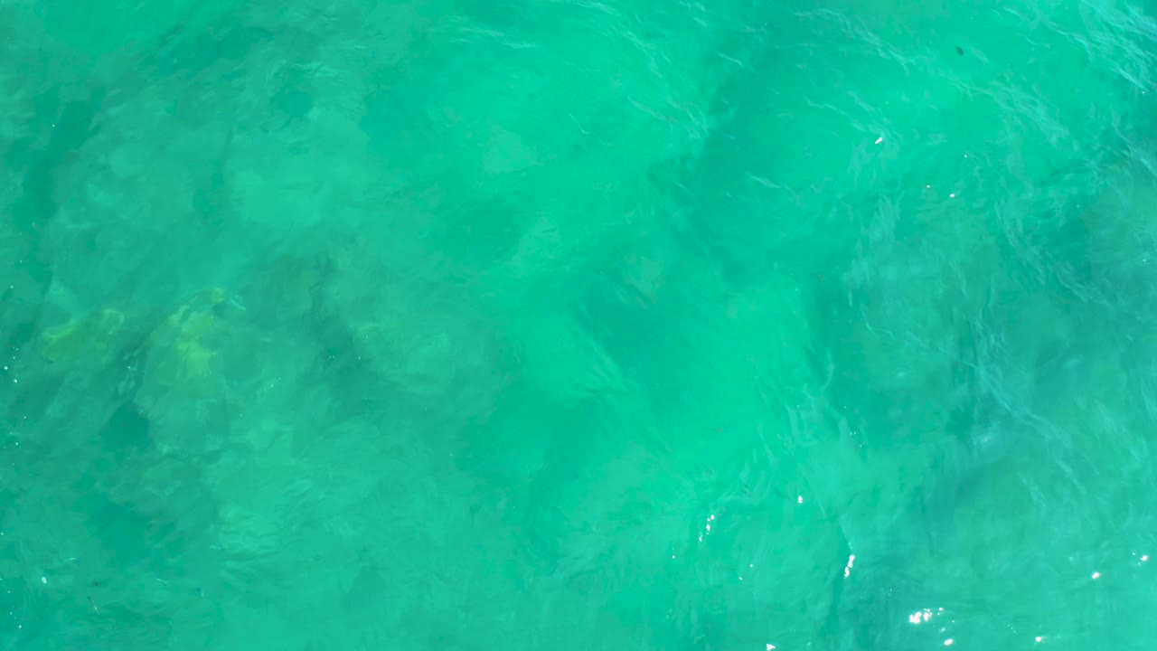 Clear blue-green ocean surface at Shipwrecks Beach, with darker underwater patches shifting under small sun highlights and no visible people.