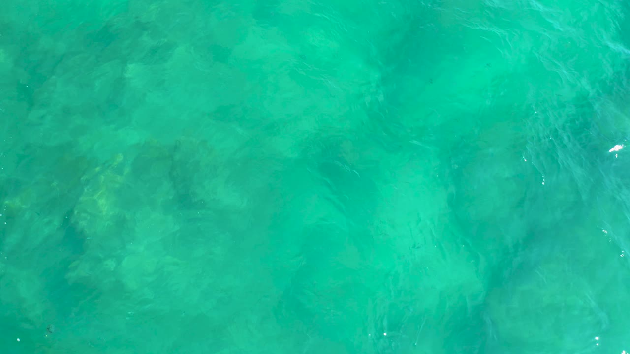 Aqua water at Shipwrecks Beach shows muted rock shapes below the surface, soft ripple lines, and no visible people or vessels.