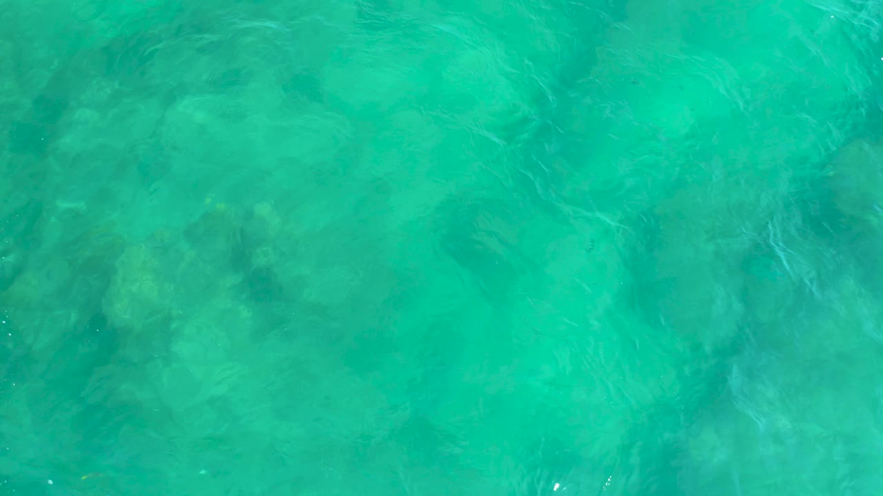 Turquoise overhead ocean view at Shipwrecks Beach, with shadowy submerged forms softened by surface ripples and no visible people.